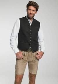 Mann, der traditionelle beige lederne bestickte Lederhosen mit einem weißen Hemd und einer dunklen, geknöpften Weste trägt, mit den Händen in den Taschen, leicht lächelnd.