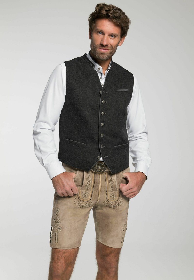 Mann, der traditionelle beige lederne bestickte Lederhosen mit einem weißen Hemd und einer dunklen, geknöpften Weste trägt, mit den Händen in den Taschen, leicht lächelnd.