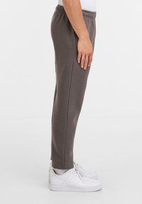 Person med grå elastiske joggers, hvide sokker og hvide Nike sneakers, stående sidelæns mod en ensfarvet hvid baggrund.