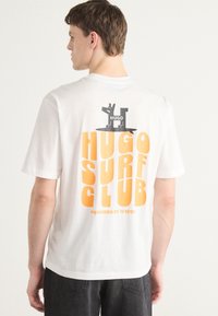 T-shirt bianca in cotone con una stampa grafica arancione audace che riporta la scritta "HUGO SURF CLUB" e un piccolo logo nero sul retro.