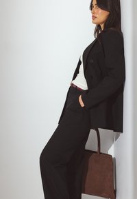 Frau im schwarzen Anzug mit weißem Oberteil lehnt an einer Wand, hält eine braune Leder-Umhängetasche und schaut seitlich.