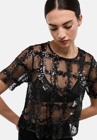 Haut noir transparent à manches courtes, orné de sequins noirs disposés en quadrillage, laissant apparaître une sous-couche avec un col en V.