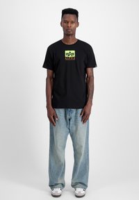 Mustriga must T-särk, millel on neoonkollane ja -oranž "ALPHA INDUSTRIES" logo, koos helesiniste vabade teksade ja valgete tossudega.