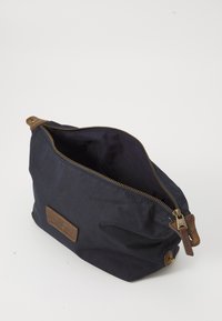 Pochette in tessuto blu navy con zip in pelle marrone e patch, parzialmente aperta, che mostra l'interno vuoto su sfondo bianco.