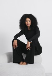 Melna garām piedurknēm topi un plašu kāju bikses, sēžot uz teksturētas baltas izliekta dīvāna, kas demonstrē mūsdienīgu, minimālistisku dizainu.