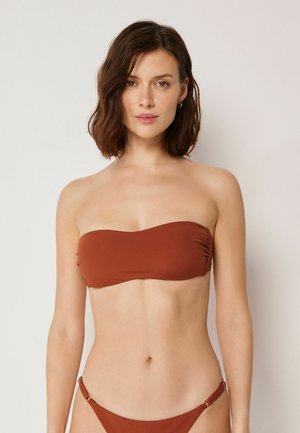 Frau mit kurzen braunen Haaren, die ein trägerloses Bikinioberteil in Rostfarbe und passende Unterteile trägt, steht vor einem einfachen hellen Hintergrund.