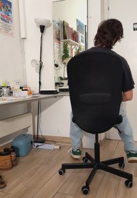 Black ergonomic office chair with a mesh back, five wheels, and adjustable height. Light wooden floor and various items visible in the room.