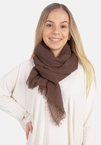 Écharpe tissée marron avec une texture douce, présentant des bords effilochés. Exposée drapée autour du cou sur un haut de couleur claire.