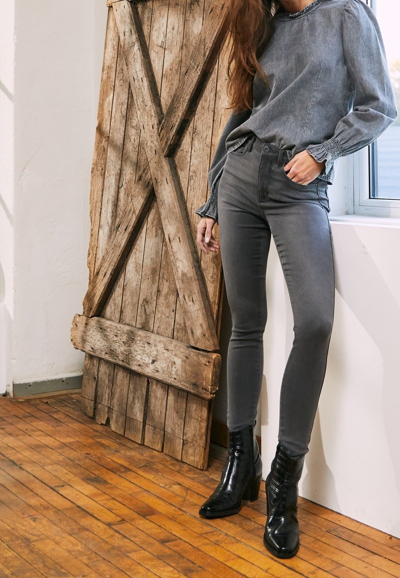 Jeans grises ajustados combinados con una blusa gris con mangas texturizadas y botas negras hasta el tobillo. El fondo incluye una puerta de madera rústica.