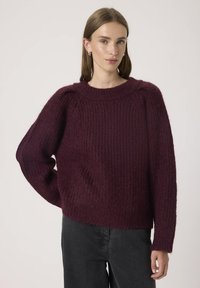 Maglione lavorato a maglia color bordeaux con una finitura testurizzata, motivo a coste e maniche ampie. Collo a giro e vestibilità rilassata. Abbinato a jeans scuri.