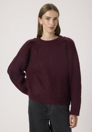Pull en maille bordeaux avec une finition texturée, motif côtelé et manches larges. Encolure ronde et coupe décontractée. Associé à un jean foncé.