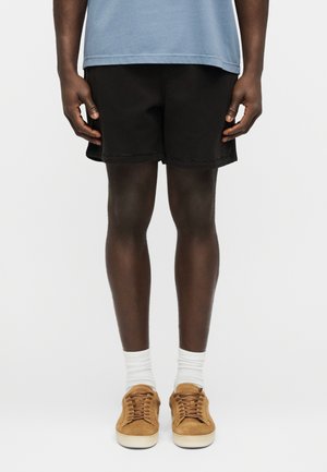 Man wearing light blue shirt, black shorts, white socks, and tan suede sneakers standing against white background.