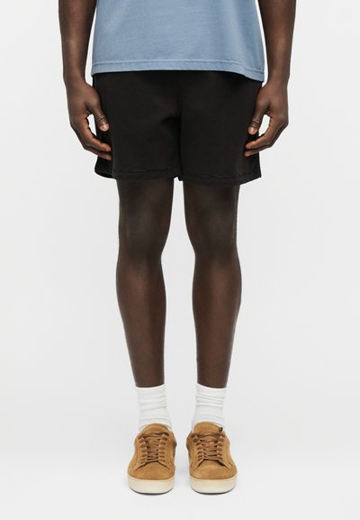 Homme portant une chemise bleu clair, un short noir, des chaussettes blanches et des baskets en suède beige, debout devant un fond blanc.