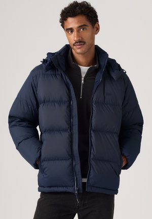 Homme portant une doudoune bleu marine par-dessus un pull noir à demi-fermeture éclair, les mains dans les poches de la veste, sur un fond uni.