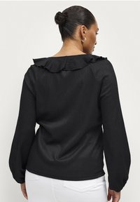 Blusa nera a maniche lunghe con tessuto testurizzato, caratterizzata da un colletto arricciato e polsini elasticizzati. La vista posteriore mostra una vestibilità rilassata.