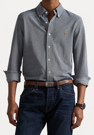 Mężczyzna ubrany w szarą koszulę na guziki z podwiniętymi rękawami, ciemnoniebieskie dżinsy, brązowy skórzany pasek i zegarek na rękę z brązowym paskiem.