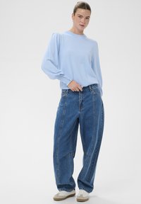 Pull bleu clair à manches longues avec épaules bouffantes, associé à un jean bleu à jambes larges avec des panneaux contrastants, et des baskets blanches.