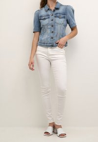 Giacca in denim azzurro chiaro corta con maniche a sbuffo, due tasche sul petto e chiusura a bottoni, abbinata a pantaloni bianchi aderenti e mule bianchi.