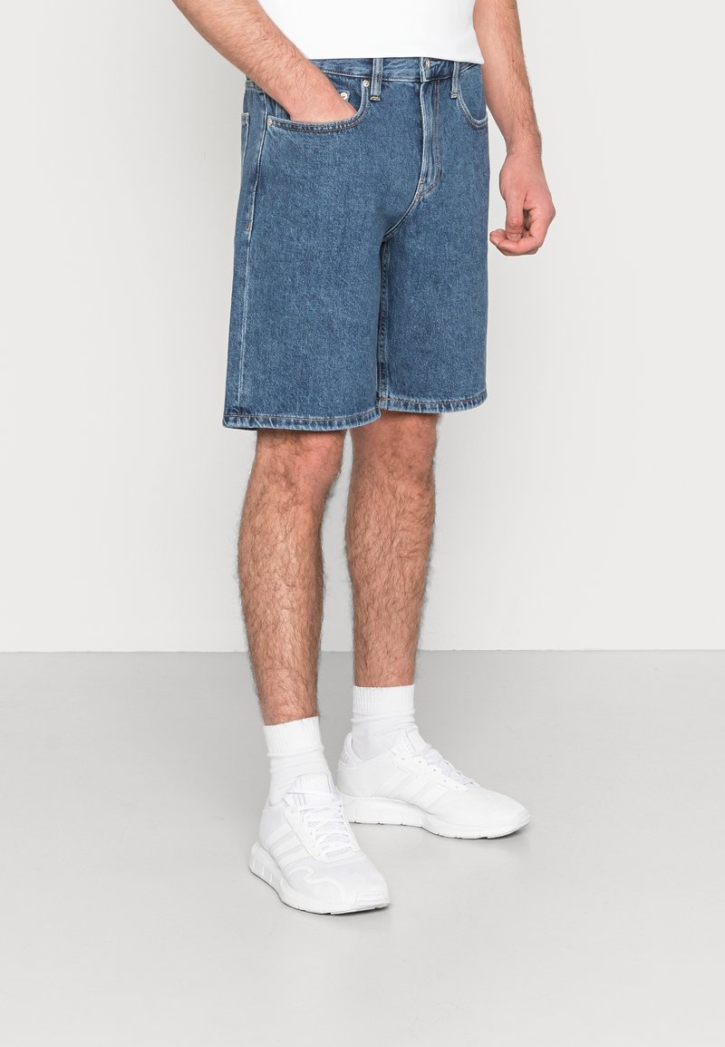Man med blå denimshorts, vita sportskor och vita strumpor som står på ett ljust grått golv mot en vit vägg.