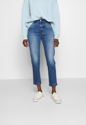 Osoba ubrana w niebieskie spodnie jeansowe do połowy łydki, jasnoniebieską bluzę i białe trampki na sznurowadła, stojąca na tle jednolitego jasnego tła.