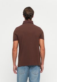 Mann med brun kortermet polo-skjorte og blå jeans står vendt mot en hvit vegg, og viser bakfra fra hodet til hoftene.