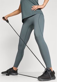 Sportos öltözet, amelyhez egy laza türkiz felső és egy testhezálló türkiz leggings tartozik. A megjelenést fekete sportcipők és egy ellenállásgumi egészíti ki.