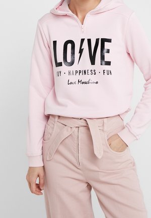 Osoba w jasnoróżowej bluzie z napisem „LOVE” oraz „JOY • HAPPINESS • FUN” i jasnoróżowych spodniach z wysokim stanem i wiązanym paskiem.