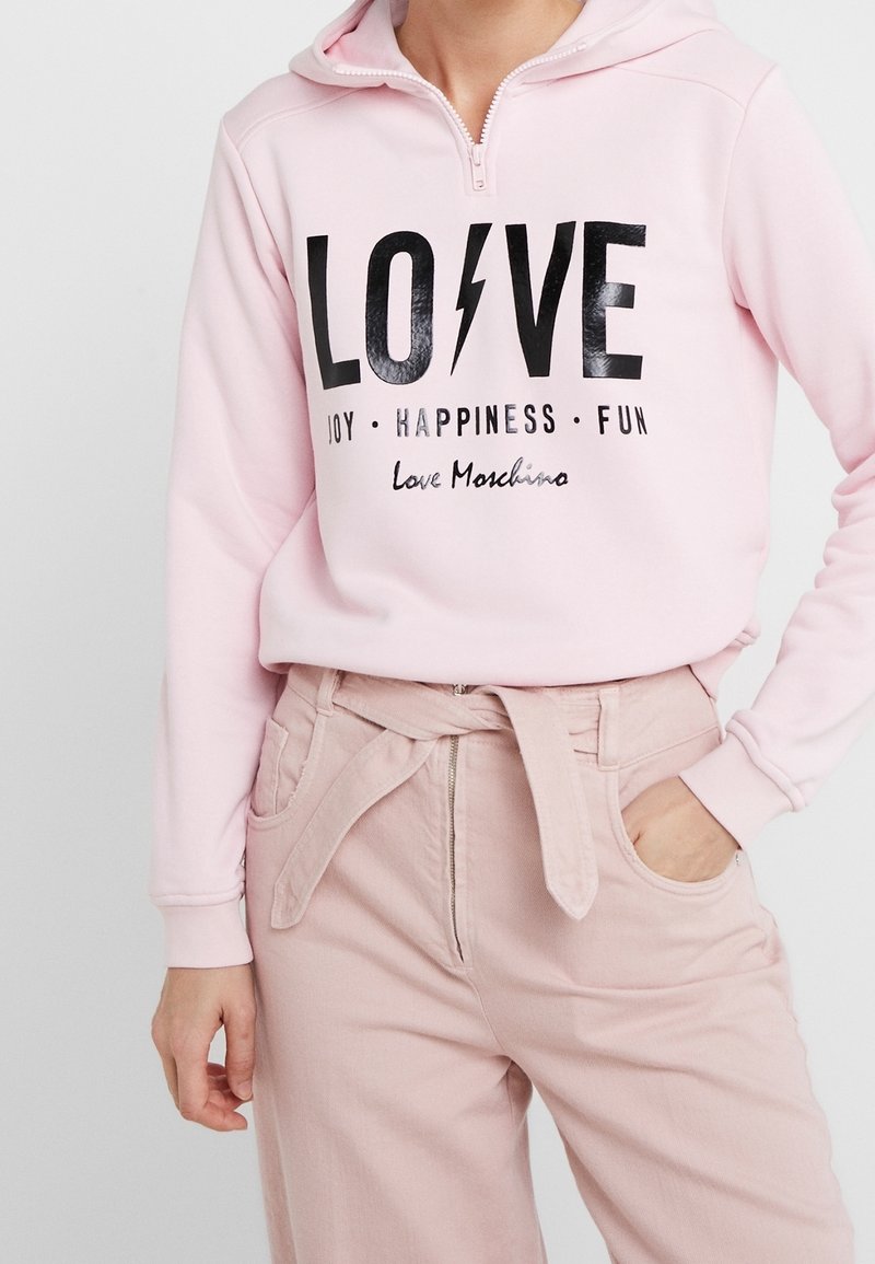 Personne portant un sweat à capuche rose clair avec les inscriptions « LOVE » et « JOIE • BONHEUR • PLAISIR » ainsi qu'un pantalon taille haute rose clair avec ceinture nouée.