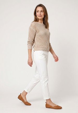Kvinde går iført en beige strikket sweater, hvide op-rullede bukser og brune teksturerede flade sko mod en ensartet lys baggrund.
