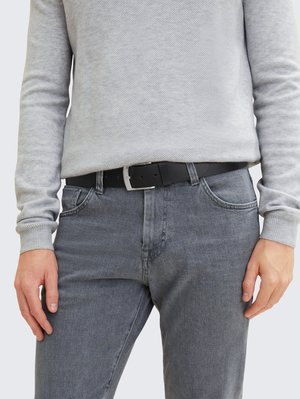 Osoba u svijetlosivom teksturiranom džemperu zataknutom u sive traperice s crnim kožnim remenom i srebrnom kopčom. Ruke opuštene uz tijelo.