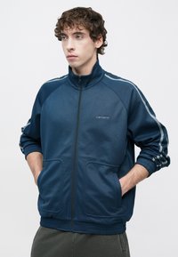 Μπλε ναυτικό τζάκετ από απαλό ύφασμα, με φερμουάρ πλήρους μήκους, ψηλό γιακά, ελαστικά μανσέτες και δύο μπροστινές τσέπες με αντίθετες ρίγες.