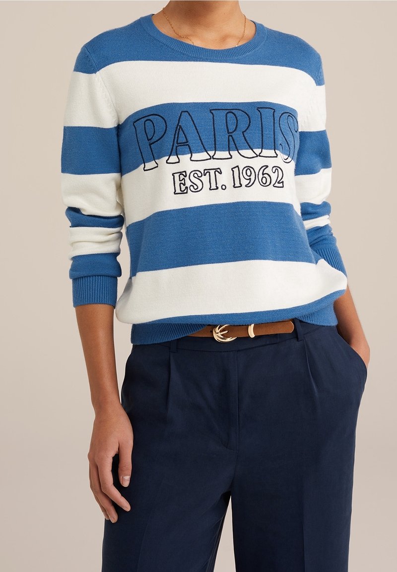 Personne portant un pull à rayures bleues et blanches avec le texte « PARIS EST. 1962 », un pantalon bleu marine et une ceinture marron avec une boucle dorée.