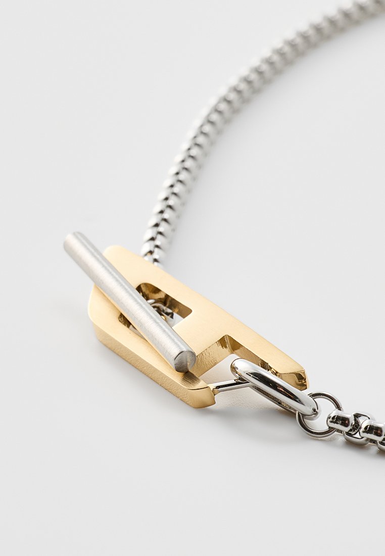 Collier en métal avec une chaîne argentée et un pendentif géométrique en or avec un accent cylindrique en argent. Surface lisse et finition polie.