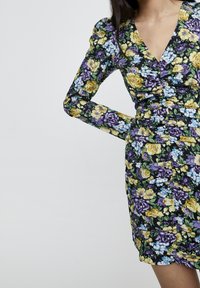 Abito floreale con maniche lunghe, scollatura a V profonda e dettagli arricciati. Il tessuto presenta vivaci fiori gialli, viola e blu su uno sfondo nero.