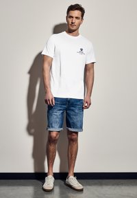 Bílý bavlněný tričko s modrým grafickým logem, doplněné odhalenými džínovými kraťasy. Model má na sobě bílé tenisky s béžovými akcenty. Neutrální pozadí.