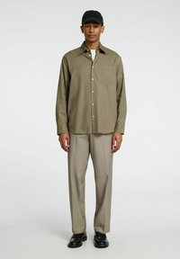 Camicia a maniche lunghe verde oliva con chiusura a bottoni, tasca singola; abbinata a pantaloni grigi chiari su misura e scarpe di pelle nera.