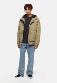 Beige huva jacka med två framfickor, kombinerad med en svart t-shirt och ljusblå jeans. Svarta slip-on skor fulländar looken.