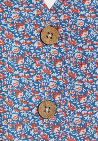 Tissu à motif floral en bleu, rouge et blanc. Présente un col en V et un bouton rond marron clair à quatre trous.