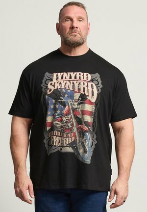 Mann trägt schwarzes Lynyrd Skynyrd T-Shirt mit Motorrad- und amerikanischer Flaggen-Grafik, steht vor schlichtem hellem Hintergrund.