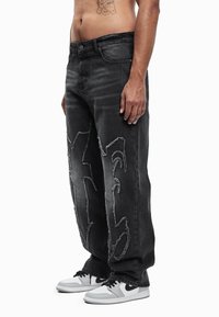 Μαύρο τζιν από denim με σκισίματα και μεγάλες εγκοπές μπροστά, σε συνδυασμό με λευκά αθλητικά παπούτσια με μαύρες λεπτομέρειες.