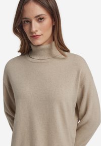 Maglione a collo alto beige realizzato in tessuto morbido. Presenta un collo a costine e una vestibilità morbida con spalle scese, che enfatizzano il comfort.