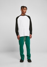 Weißes Langarmhemd mit schwarzen Raglanärmeln, kombiniert mit grünen Sweatpants. Verfügt über einen Kordelzugbund und schwarze Sneakers.