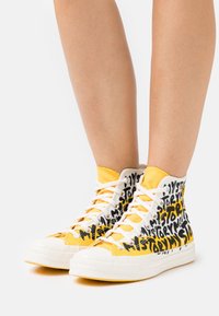 Vita högskor i sneakersstil med svarta och gula textmönster. Kontrasterande gul accent upptill med vita snören och gummisula.