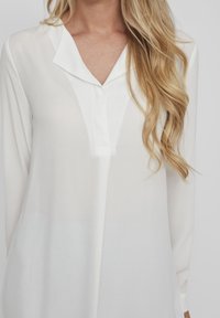 Vit blus med långa ärmar, V-ringning och knapppdetalj, tillverkad av lätt och genomskinlig tyg. Texturen ser slät och något genomskinlig ut.