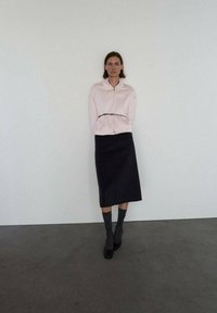 Blouse zippée rose clair avec col, associée à une jupe noire mi-longue. Le modèle porte des chaussettes foncées et des chaussures noires. Textures lisses tout au long.