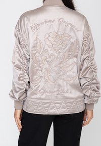 Femme portant une veste bomber en satin beige clair avec manches froncées, motif floral brodé et texte dans le dos.