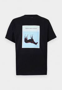Μαύρο Τ-shirt με μια κεντρική, ανάποδη καφέ εικόνα αλόγου μέσα σε μπλε τετράγωνο και το κείμενο "UPSIDE DOWN HORSE" πάνω από αυτή.