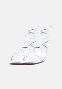 Ζευγάρι ασημένια σανδάλια με λουριά και ψηλό τακούνι, με λουριά που τυλίγονται γύρω από τον αστράγαλο και τετράγωνη μύτη, σε λευκό φόντο.