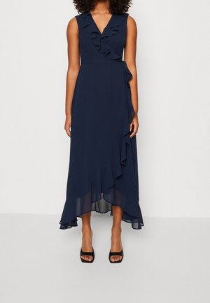 Robe portefeuille sans manches bleu marine avec bordure à volants, col en V, ourlet asymétrique, portée avec des talons ouverts noirs.