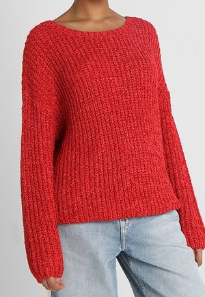 Pull en grosse maille rouge à manches longues porté avec un jean bleu clair, montré du milieu du cou jusqu'aux hanches sur une personne.